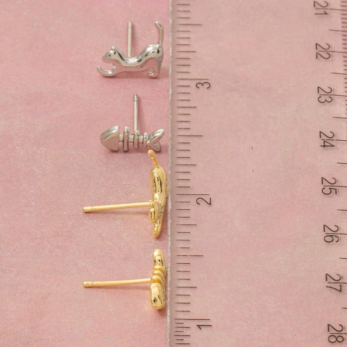 Silver cat and fish shaped stud earrings on a pink background with ruler for scale