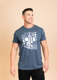 Man wearing Fading Saguaro Tee in blue heather with a white graphic, posing against a beige backdrop.