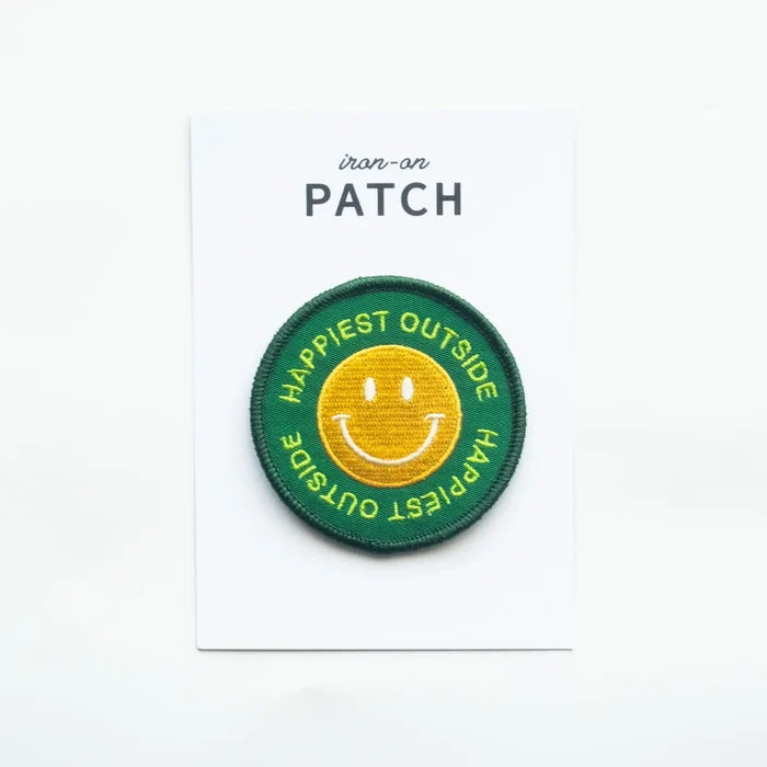 Close up of round Happiest Outside patch with green border and yellow smiley on a white display card