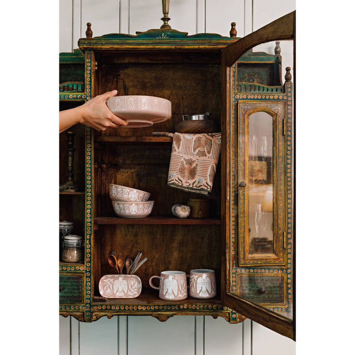 Hand holds a pale pink Peregrine Imprint Mug with a white pattern, about to be placed in a vintage cabinet with other dishware.