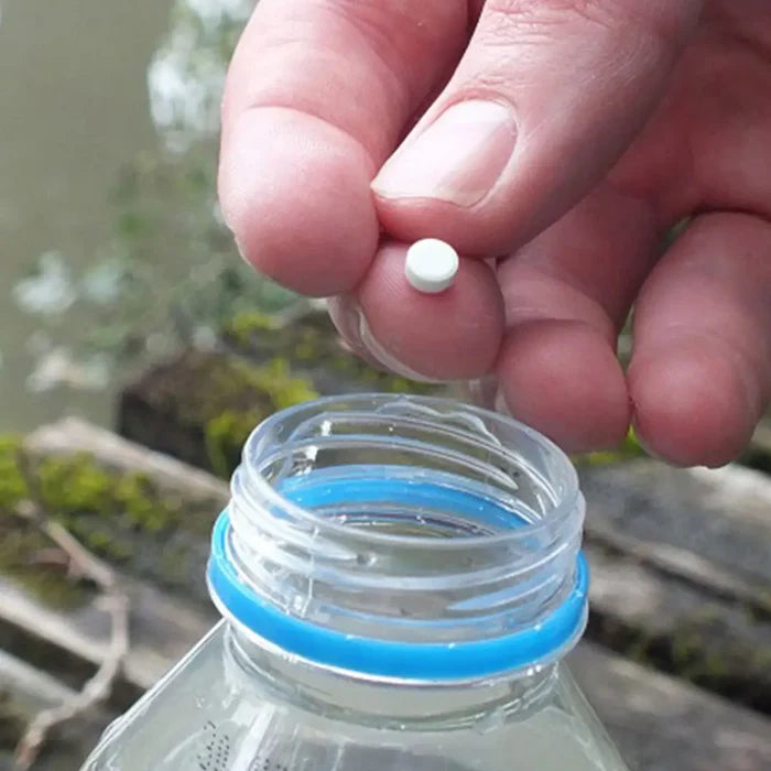 Hand holding a Pyramid Biox Aqua Chlorine Dioxide Water Purification Tablet above a clear plastic bottle with a blue cap, outdoors.