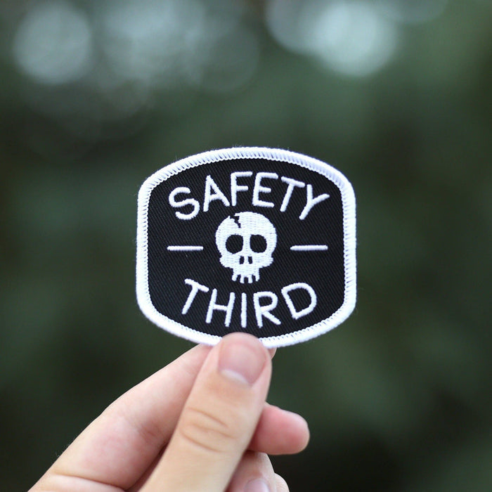 Patch held by fingers showing a skull and the Safety Third design against a blurred green background