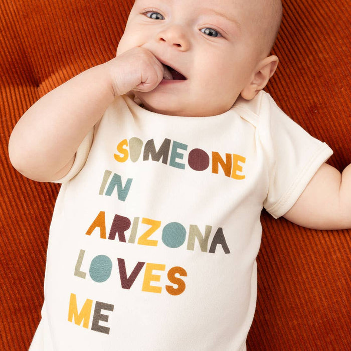 Baby wearing a white onesie with colorful text that reads SOMEONE IN ARIZONA LOVES ME