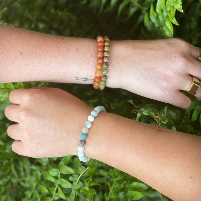 Two arms outdoors wearing unakite bead bracelets in green and coral tones along with a pale blue bracelet.