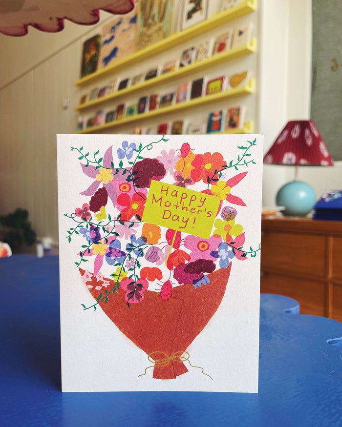 Vibrant bouquet card held up in a bright room with shelves and a red lampshade in the background