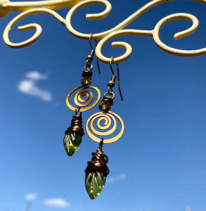 Brass spiral olive leaf earrings hanging from a scrollwork stand against a clear blue sky