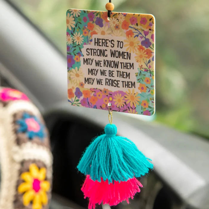 Floral patterned car air freshener hanging from a rearview mirror with turquoise and pink tassels and the message Here's to strong women