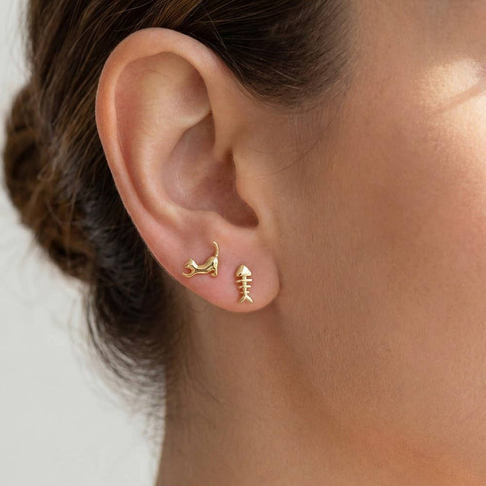 Gold cat and fish stud earrings on the ear featuring a leaping cat and a fish skeleton