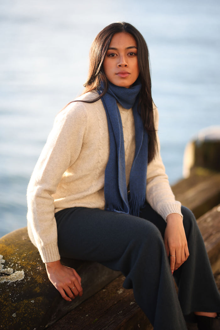 Woman wearing a cream sweater and blue scarf sitting by a body of water.