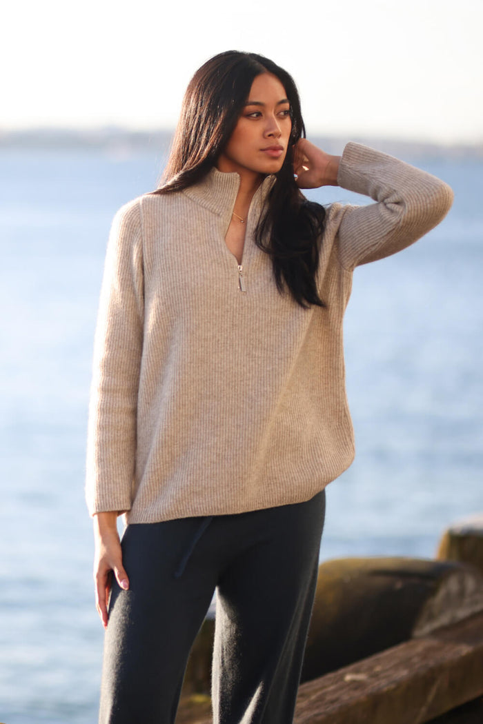 Woman wearing a beige sweater and black pants standing by the water.