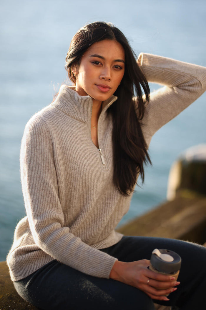Woman in a beige sweater holding a coffee cup by the water