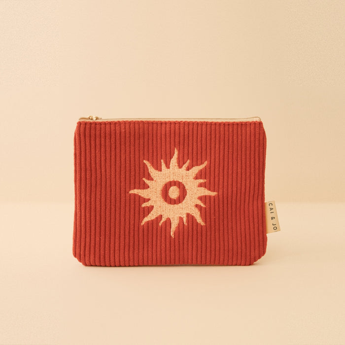 Rust corduroy pouch with a white sunburst embroidery and a fabric tag on the side.