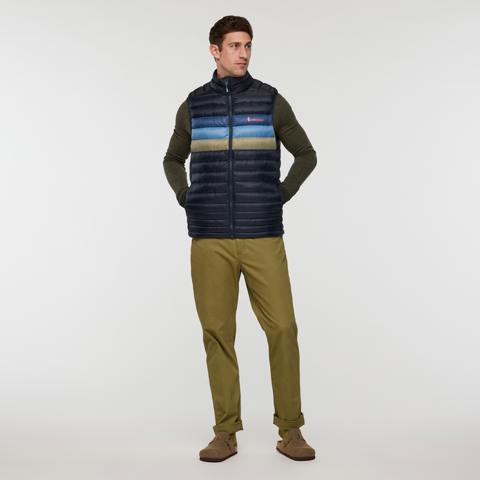 Man wearing Cotopaxi Fuego Down Vest in dark navy with blue and tan stripes over olive sweater and khaki pants