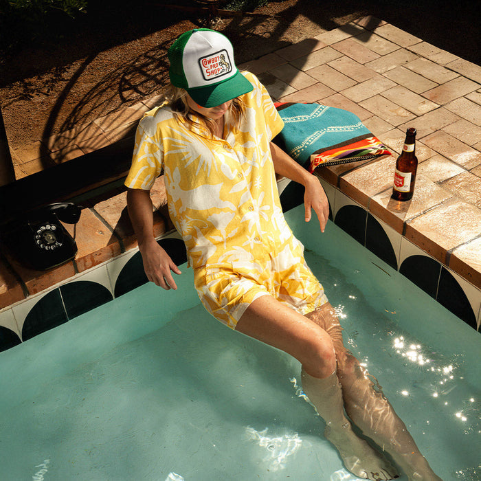 Person lounging by a pool wearing a yellow tropical shirt and shorts, sporting a green and white mesh Cowboy Pro Shop hat.