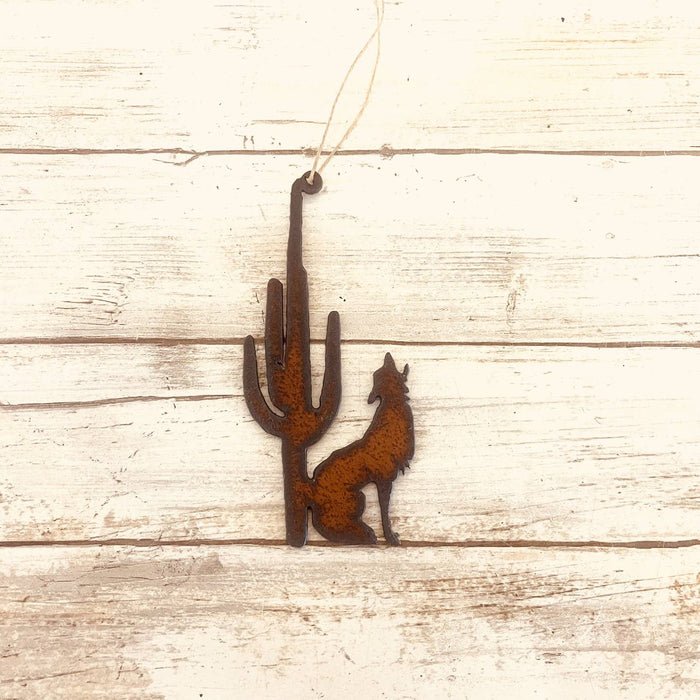 Rusty metal ornament showing a coyote beside a tall cactus hanging on twine against a whitewashed wooden background
