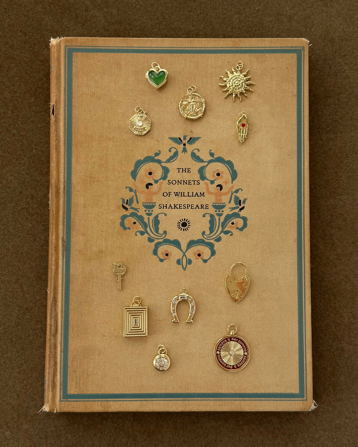 A vintage book titled "The Sonnets of William Shakespeare" rests on a brown surface. Twelve charms, including the Mod + Jo Davie Heart Birthstone Locket Charm with genuine gemstones, are arranged around the ornate cover.