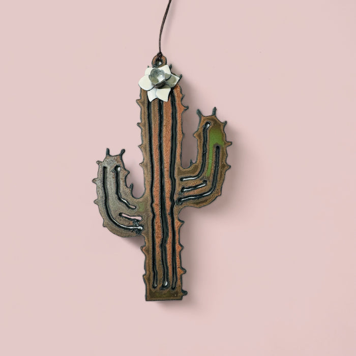 Desert cactus flower ornament in raw metal with a white bloom on top hanging from a cord against a pink backdrop