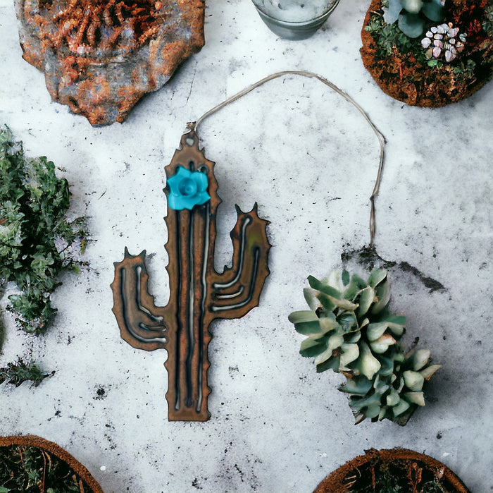 Two piece rusted metal desert cactus ornament with a turquoise flower on top laid on a concrete surface among succulents
