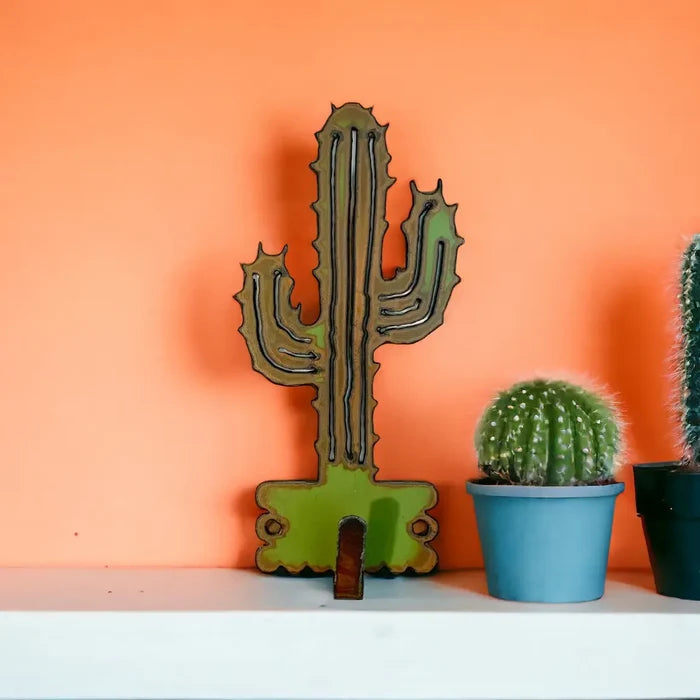 Cactus shaped wall hook in green and brown tones on an orange wall, beside small potted cacti in blue and dark pots.