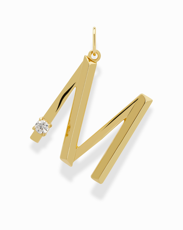 The Eloise Letter Charm by Mod + Jo is a gold pendant shaped like the letter M, accented with a sparkling cubic zirconia on the upper left side, shown against a white background.