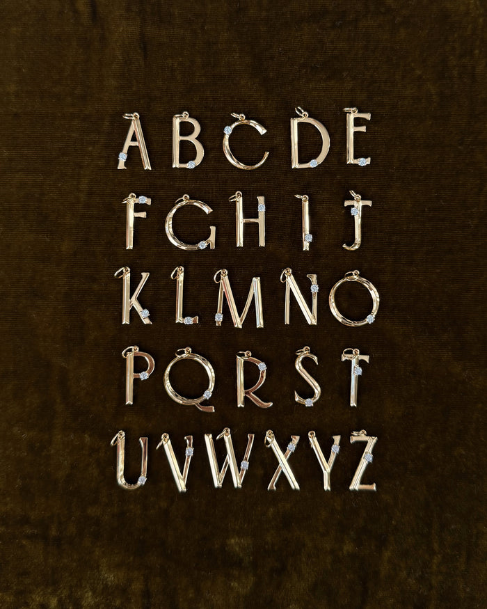 The Eloise Letter Charm by Mod + Jo features gold uppercase alphabet pendants with sparkling cubic zirconia accents, arranged in five rows on a dark brown velvet background, displaying A to Z.