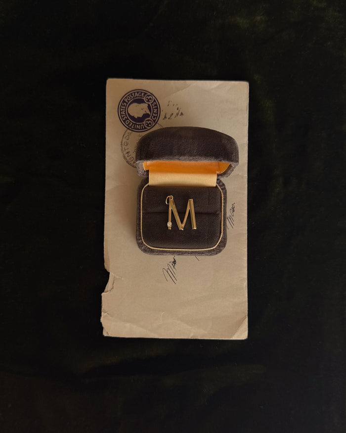 The Eloise Letter Charm by Mod + Jo, a gold "M" initial brooch with a small pearl accent, is displayed in a brown velvet box with yellow lining atop a vintage envelope featuring handwritten signatures and a postal stamp.