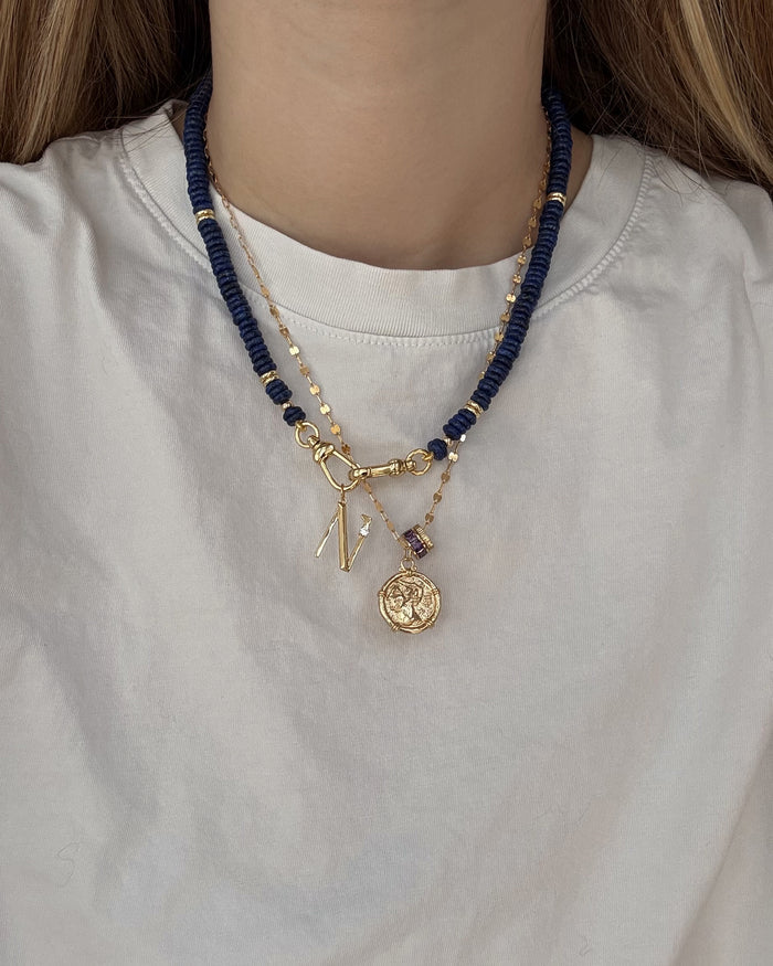 A person wears a white shirt with two layered necklaces: Mod + Jo's Eloise Letter Charm—a personalized gold initial and medallion—and a second necklace with dark blue beads and gold accents.