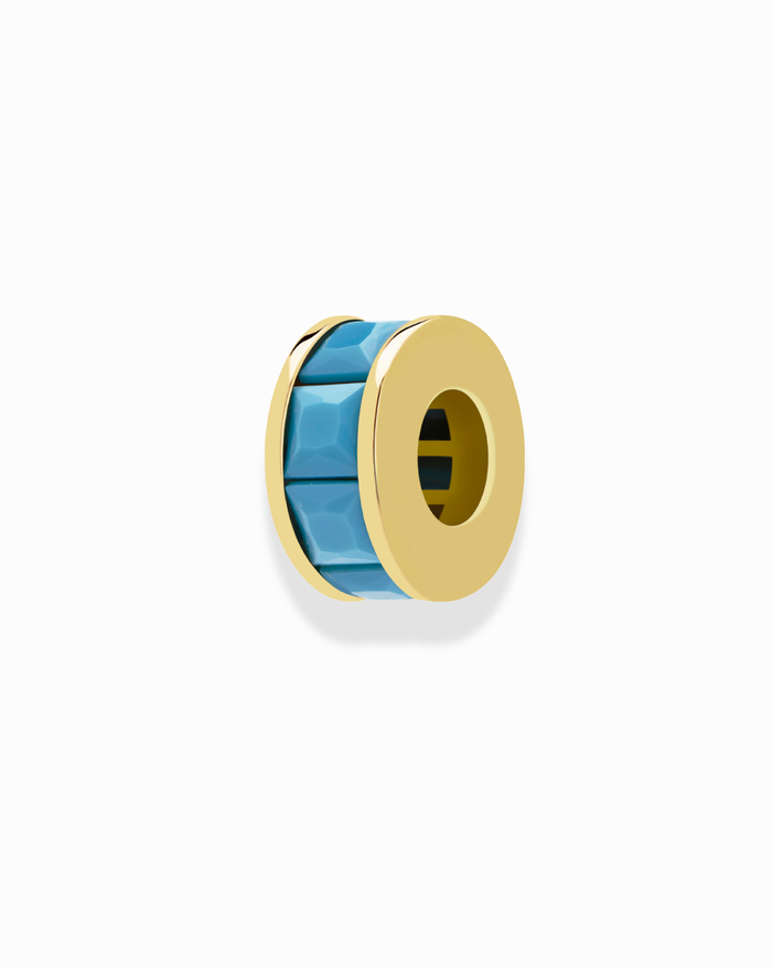 The Este Birthstone Charm Spacer by Mod + Jo features a gold circular design with faceted turquoise blue enamel accents, perfect for adding a personalized touch to your jewelry. Shown against a plain white background.