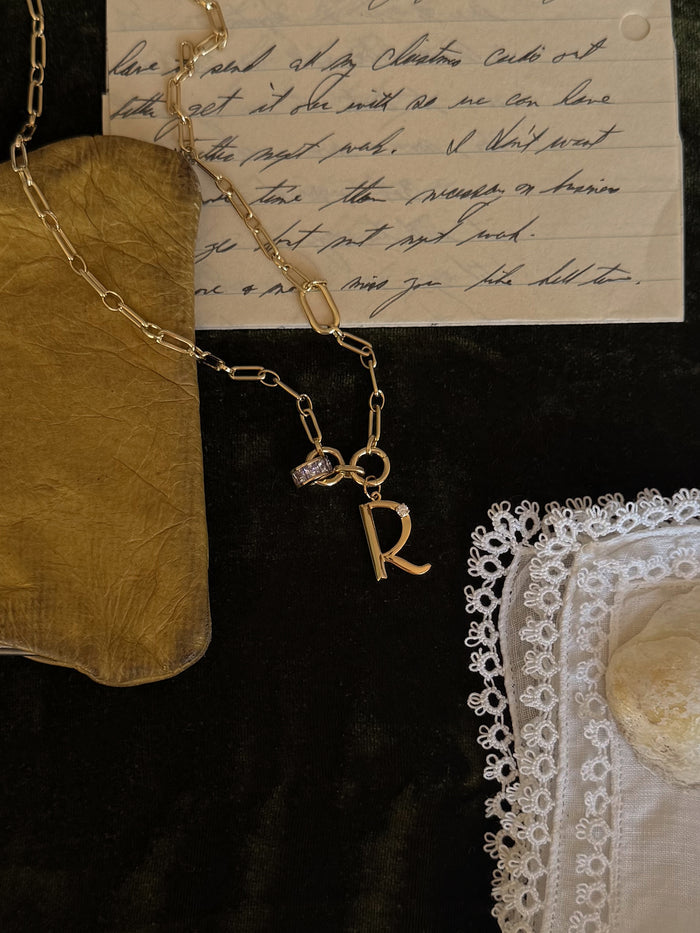 The Mod + Jo Este Birthstone Charm Spacer, a personalized 18k gold plated necklace with an R charm, rests on handwritten letters, a gold pouch, white lace-trimmed handkerchief, and a stone on dark fabric.
