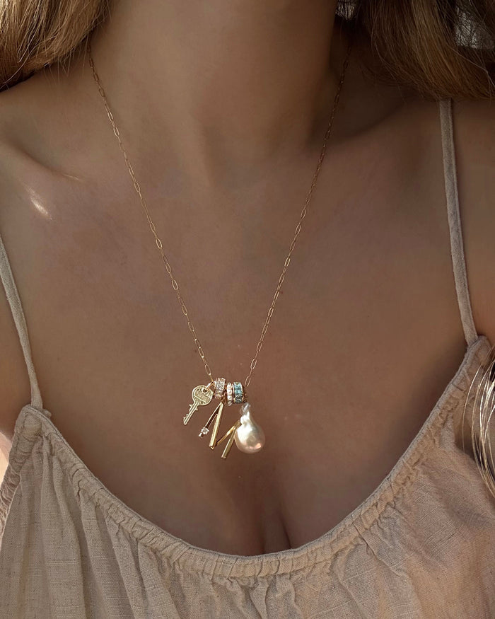A close-up of a woman wearing the Mod + Jo Eloise Letter Charm gold necklace featuring multiple pendants—key, pearl, cubic zirconia accents, and letter charms—over a beige spaghetti strap top.