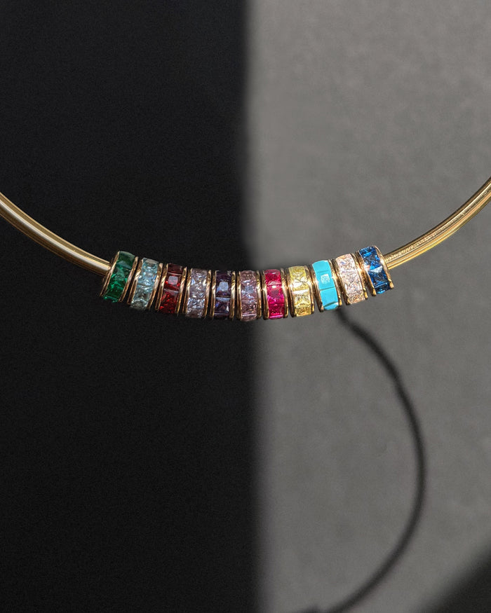 The Mod + Jo Este Birthstone Charm Spacer necklace displays nine colorful gemstone rings, each separated by a slim gold band, elegantly arranged in a row on a dark background with striking shadows.