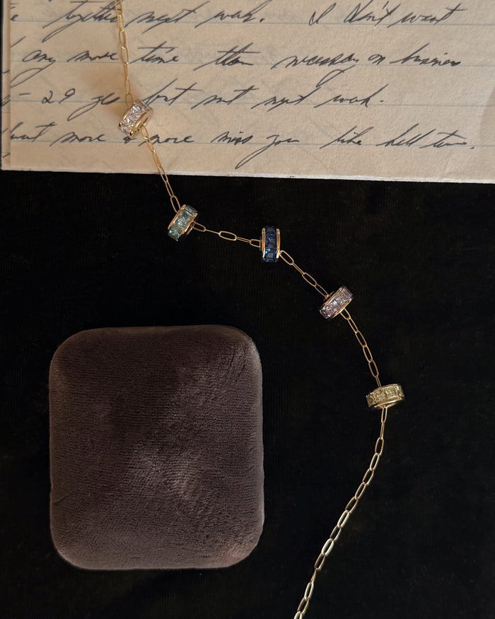 The Mod + Jo Este Birthstone Charm Spacer accents a gold chain with five jeweled rings, shown beside a closed black velvet jewelry box and a handwritten letter on lined paper.