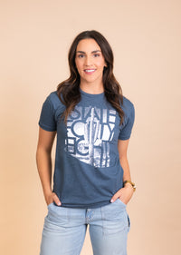 Woman wearing Fading Saguaro Tee in blue with a white graphic, standing against a beige backdrop.