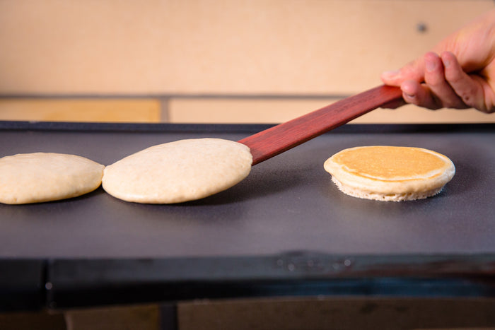 Pancakes cooking on a flat griddle as a hand uses a wooden spatula to lift one