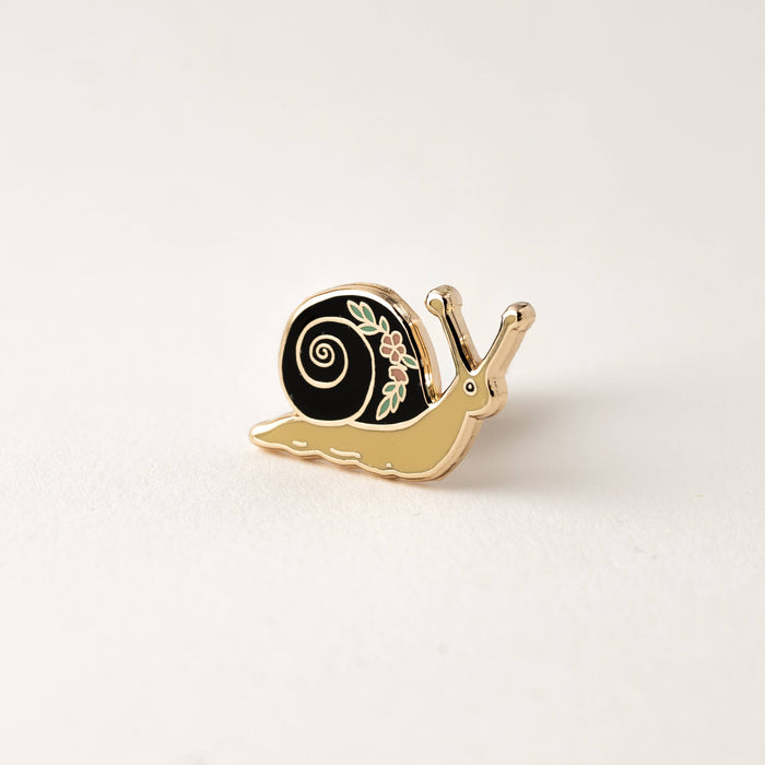 Partial view of a gold enamel garden snail pin with a black shell and floral accents resting on a light surface.