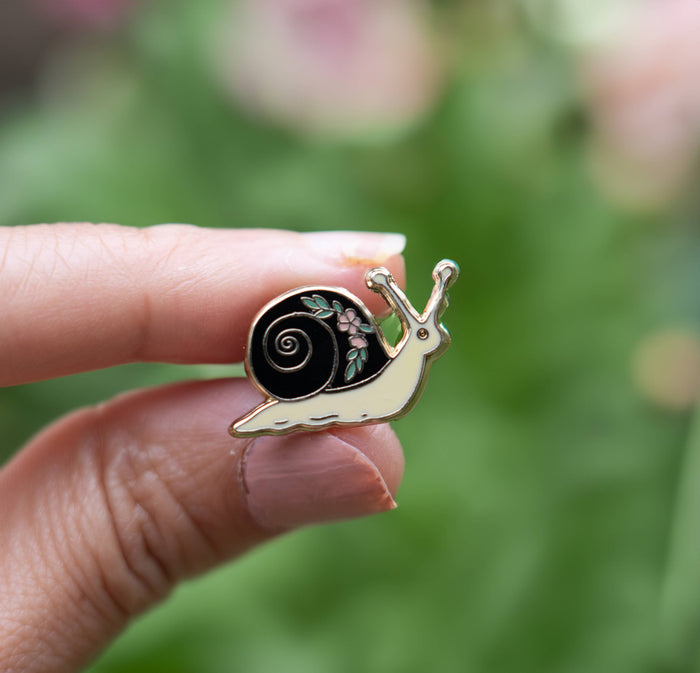 Close up of a gold enamel garden snail pin held between fingers against a blurred green background.