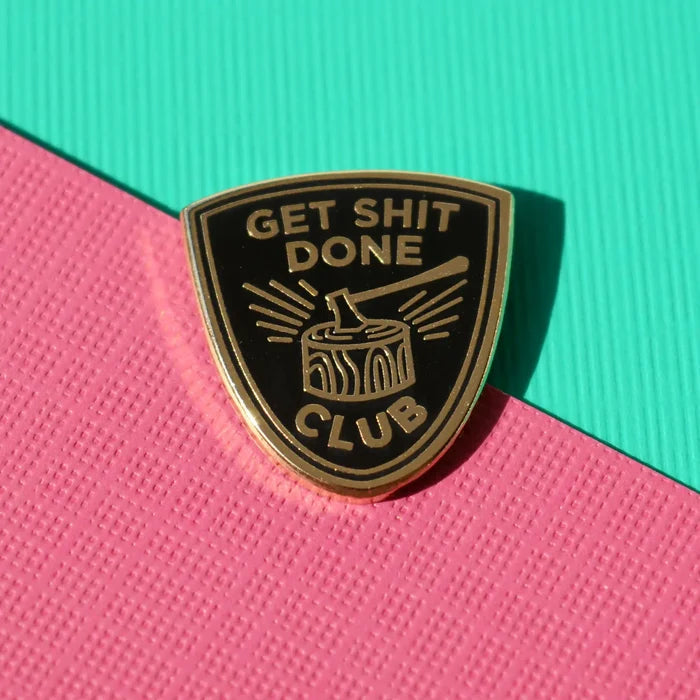 Shield shaped enamel pin with a gold edge and Get Sh*t Done on a black background.