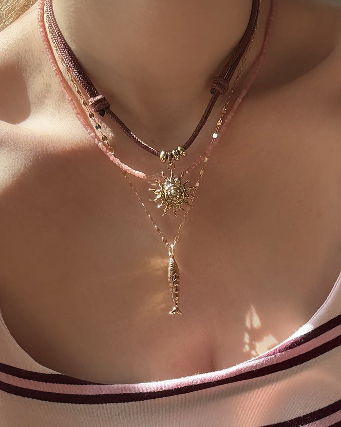 A close-up of a person’s neck wearing three layered necklaces: Mod + Jo’s Gillian Spiral Sun Charm on a brown cord with gold beads, a pink beaded choker, and an 18k gold-plated fish pendant chain. The person wears a striped top.