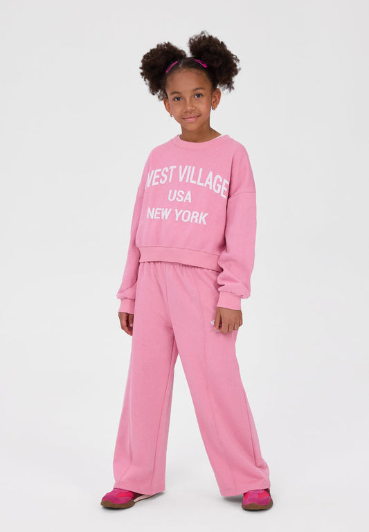 A young girl with two buns of hair wearing a pink two-piece sweatshirt and pants set with 'WEST VILLAGE USA NEW YORK' printed in white on the sweatshirt, standing against a light gray studio background.