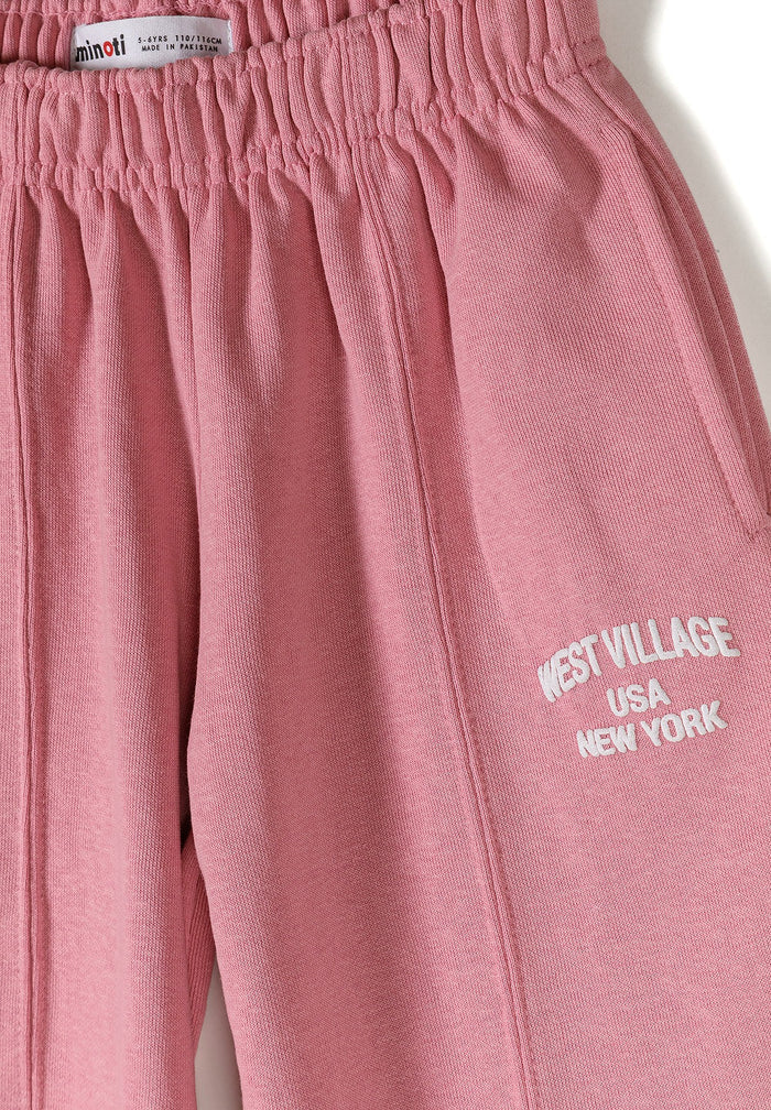 A close-up of dusty pink sweatpants featuring an elastic waistband and white embroidered text on the leg that reads 'WEST VILLAGE USA NEW YORK'.