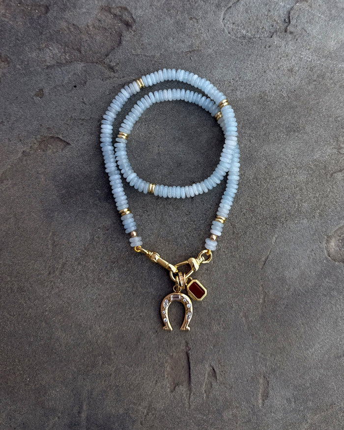 The Mod + Jo Grande Chantal Horseshoe Charm bracelet features pale blue beads, gold accents, a gold horseshoe charm with clear stones, and a red charm—shown on a textured gray surface for a touch of luck.