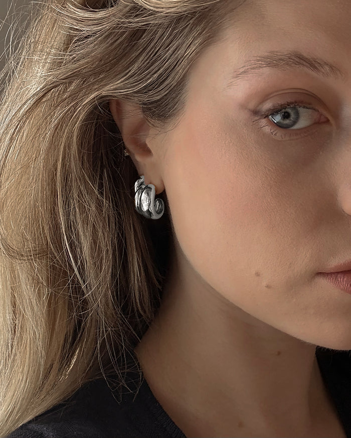 Close-up of a woman with light skin and blonde hair wearing Mod + Jo's Grande Sofie Volume Hoops. Only half her face and one blue eye are visible, softly lit to highlight the chunky rhodium plated silver earrings.