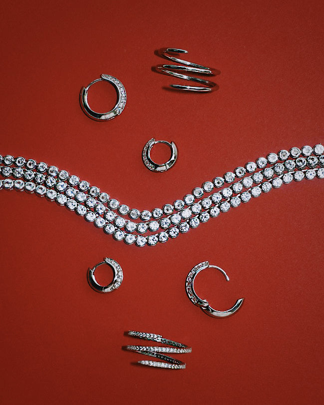 Silver hoop earrings, the Greer Pave Spiral Ring, and a three-strand diamond bracelet are arranged on a red background in a symmetrical, decorative pattern.