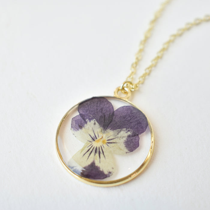 Close up of a silver chain holding a round glass pendant showcasing a purple pressed flower.