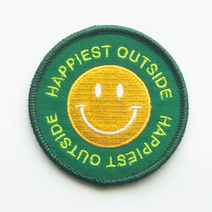 Round green embroidered patch with a yellow smiling face and lime text reading HAPPIEST OUTSIDE around the edge