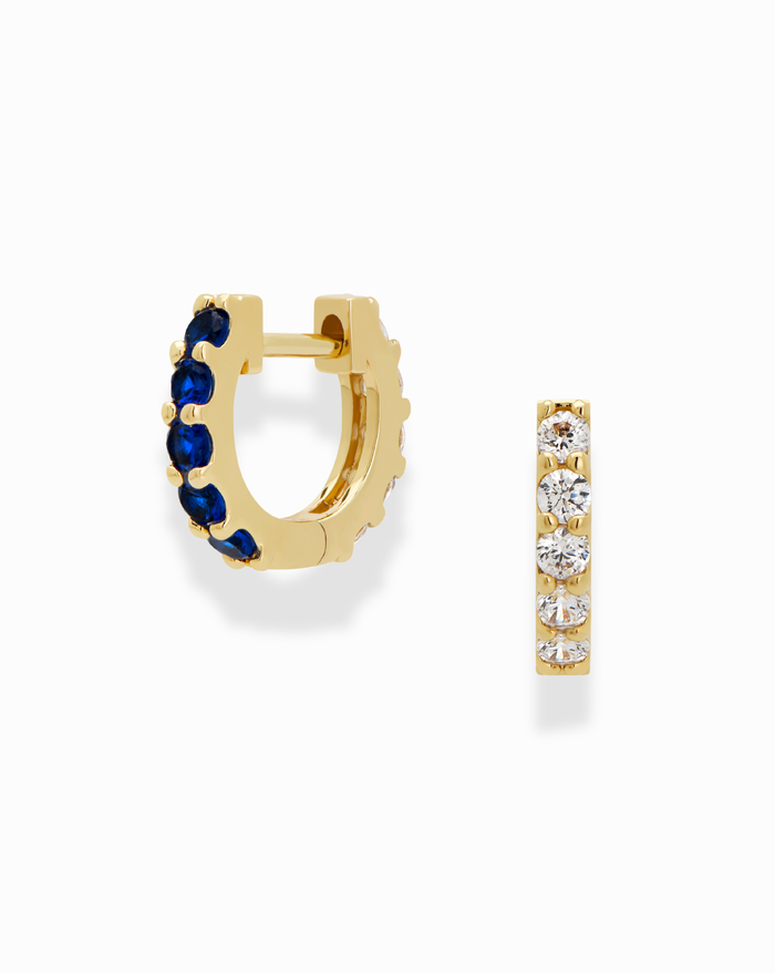 Mod + Jo's Harlow Reversible Birthstone Huggies feature small gold hoops—one side with blue birthstones, the other with clear stones—offering two elegant looks in one accessory, displayed on a white background.