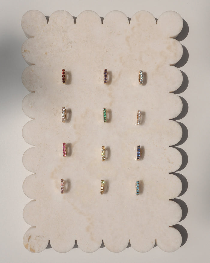 The Mod + Jo Harlow Reversible Birthstone Huggies are displayed on a scalloped-edge marble tray, with nine pairs of gold plated hoops featuring different gemstones and birthstones arranged in three rows and three columns.