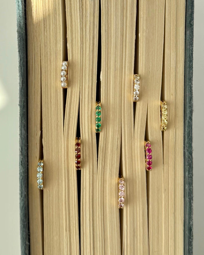Seven pairs of Mod + Jo Harlow Reversible Birthstone Huggies, each featuring a different colored gemstone, are shown inserted between the closed pages of a book, with the birthstone earrings peeking out.