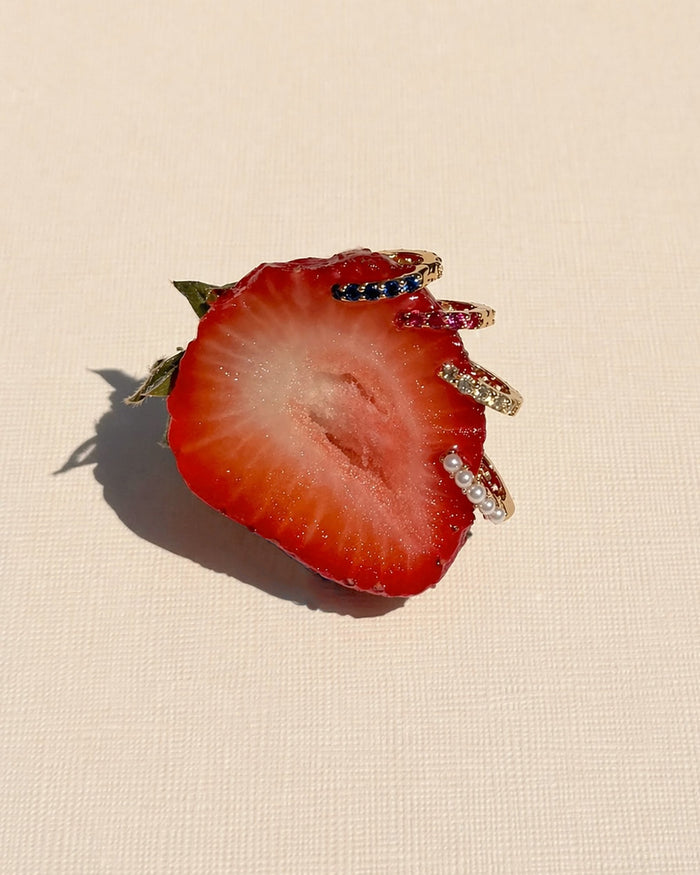 A sliced strawberry half displays the Mod + Jo Harlow Reversible Birthstone Huggies, adorned with colorful gemstones and pearls, set against a textured light background.