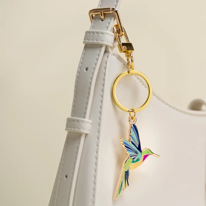 Hummingbird enamel charm on a gold key ring attached to a white bag strap with gold hardware